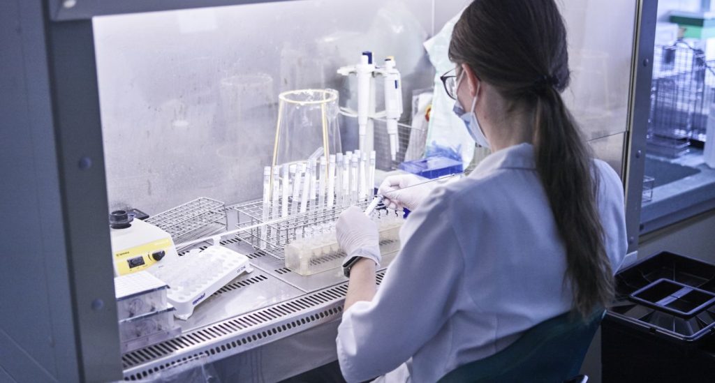 Ženska v halji obrnjena s hrbtom sede dela v laboratoriju.